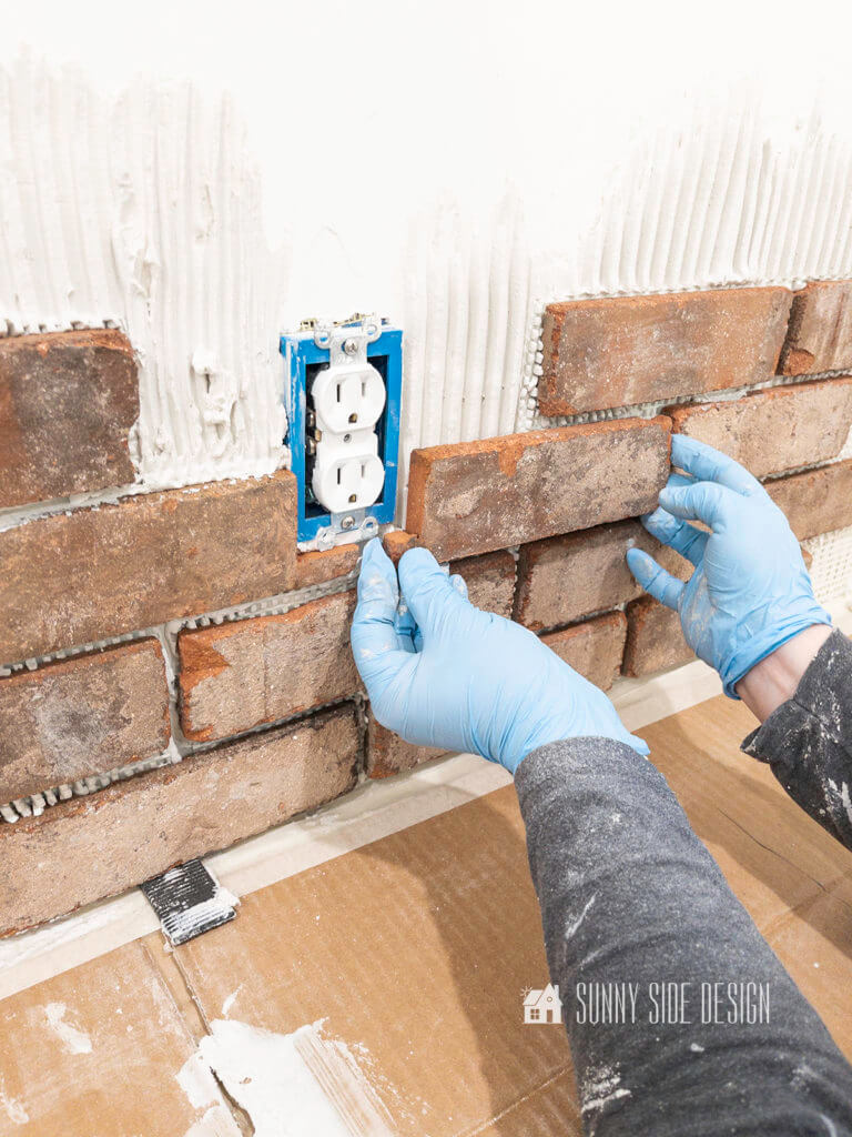 Woman's hands wearing blue latex gloves, holded cut brick veneer up to the wall to see if it will fit around the electrical outlet.