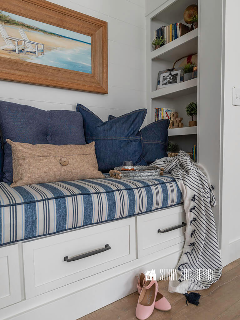 Built in reading nook in a closet with bookshelves styled with books, plants and frames. Blue striped cushion with denim and tan pillows. Framed canvas beach art on a shiplap wall. Navy and white throw blanket on cushion with a galvanized tray and a drink.