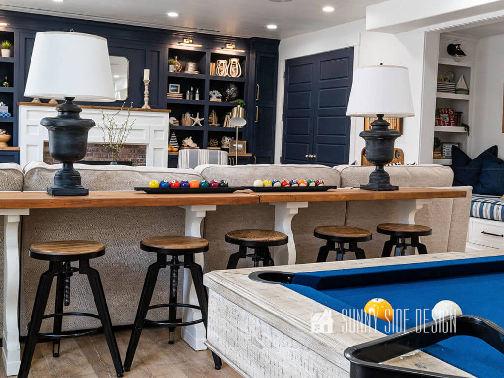 2 lamps setting on large sofa table after makeover. 5 stools at table with a centerpiece of pool table balls in wooden dough bowls, navy blue built in entertainment center in the background.