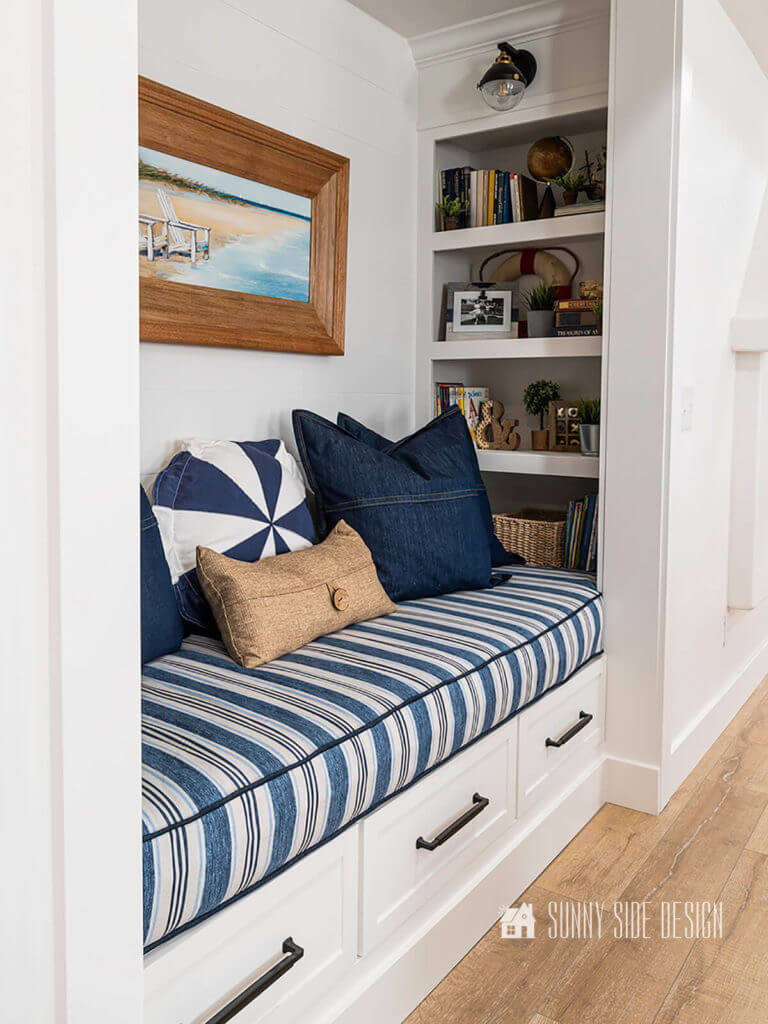 Make a box cushion, the finished cushion for the bench in the reading nook. Blue and white striped fabric with denim piping, denim pillows with a blue and white circle pillow and a sand color pillow. Coastal art on the back shiplapped wall, books and decor on the shelves.