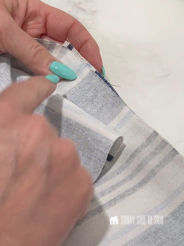 Make a box cushion, woman's hand placing a pin at the corner of the cushion, aligning the stripes of the blue and white fabric before stitching together.