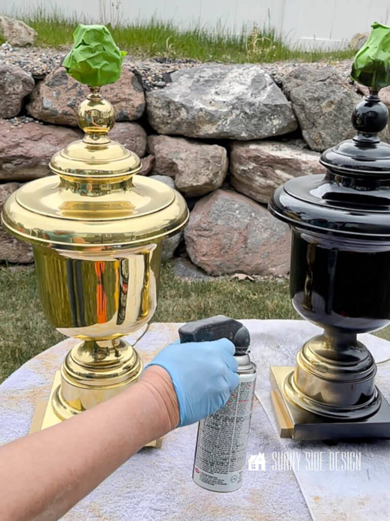 Woman's hand holding a can of black spray paint applying paint to 2 vintage brass lamps before lamp makeover