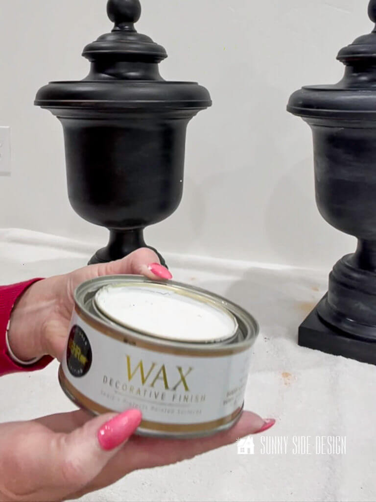 Woman's hands holding a an of white wax decorative finish in front of 2 black spray painted lamps
