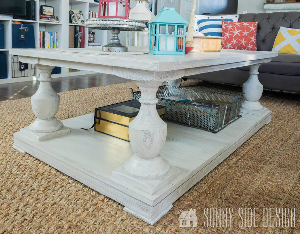 Painting furniture: Coffee table painted white with a grey glaze for a coastal look on a sisal rug, and a grey couch with blue, white, red and yellow pillows. Coffee table decorated with books, 3 lanters, and a wooden bowl.
