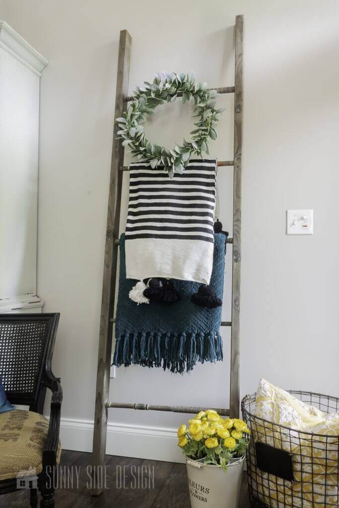 Flea market flip ideas, old wood and croquet mallets are upcycled into a blanket ladder. At the base of the ladder is a bucket with yellow flowers.