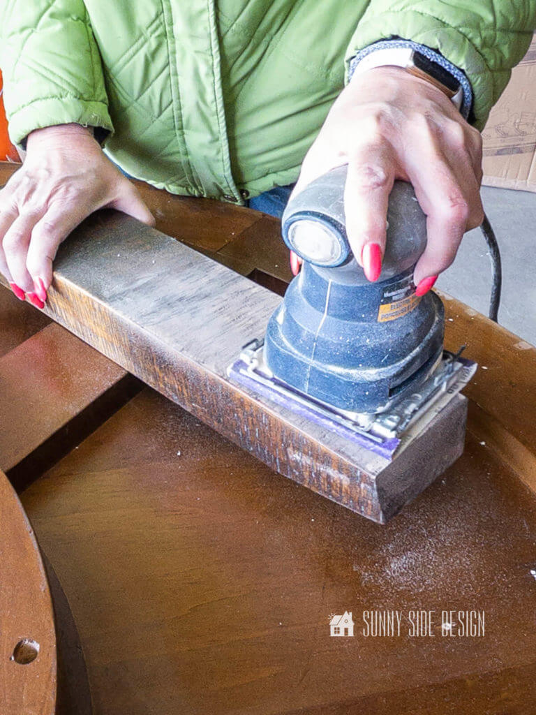 Woman's hand holding a palm sander removing the finish on the wood legs for the round coffee table.