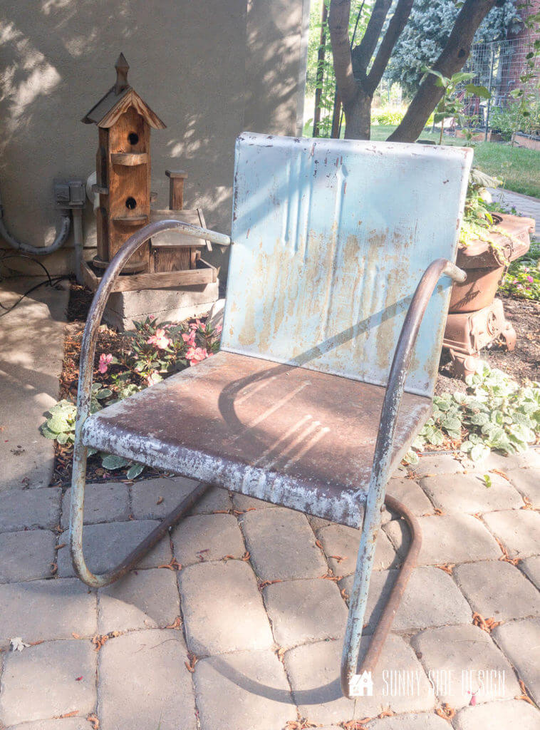 Vintage aqua blue and rusty metal chair before it's refinished, sitting in a paver patio with flowers and birdhouse in the background
