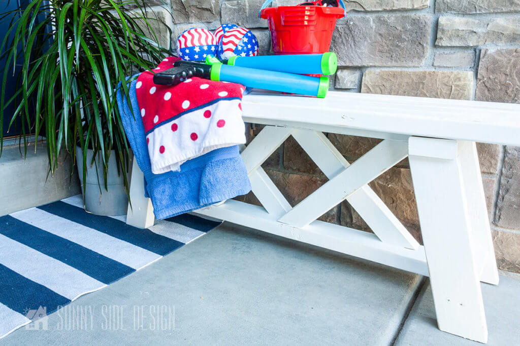 2x4 outdoor bench painted with white exterior paint, with a blue and white striped rug, plant and summer toys.