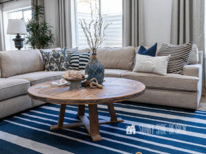 Finished Farmhouse round coffee table in the basement family room, setting on a blue and white striped rug with a sand colored sectional, blue and white decor pillows, sofa table lamps. Coffee table is decorated with a blue jug vase with branches, rope, amd a galvanized pedestal container filled with seashells.