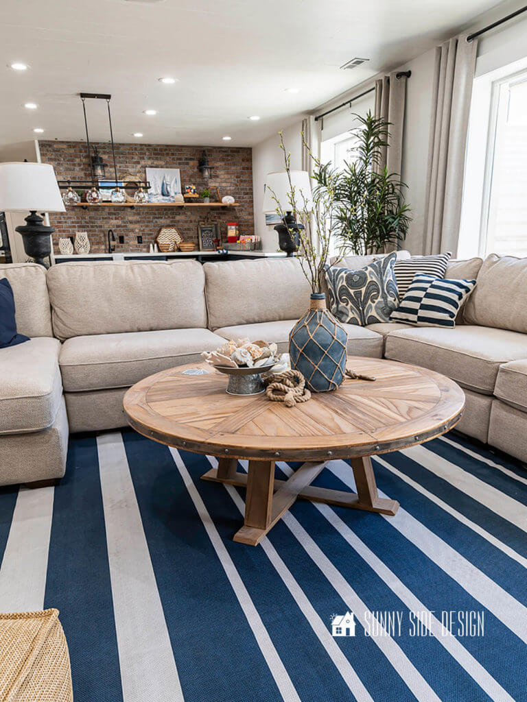 Finished Farmhouse round coffee table in the basement family room, setting on a blue and white striped rug with a sand colored sectional, blue and white decor pillows, sofa table lamps. Coffee table is decorated with a blue jug vase with branches, rope, amd a galvanized pedestal container filled with seashells. Brick wall backsplash in the background.