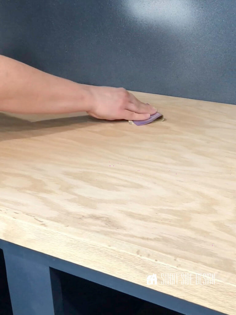 How to apply a water based stain - Woman's hand holding purple fine sandpaper lighty sanding the wood surface after the stain is applied.