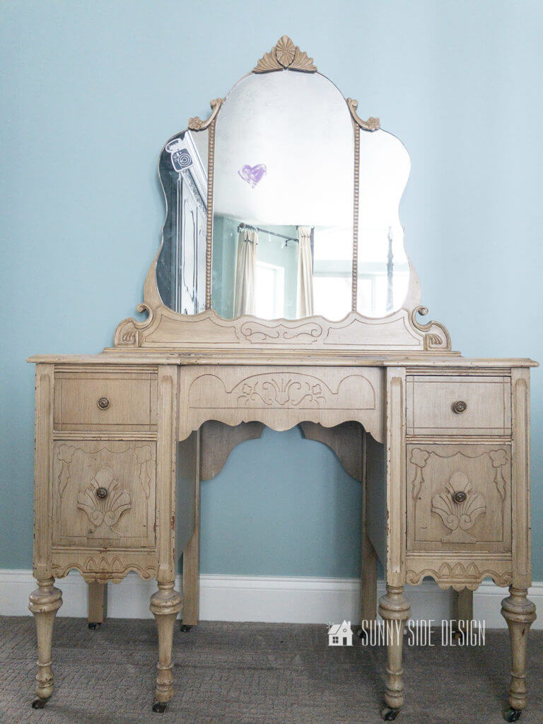 Vintage cream painted vanity with mirror before it's makeover against a light blue wall.