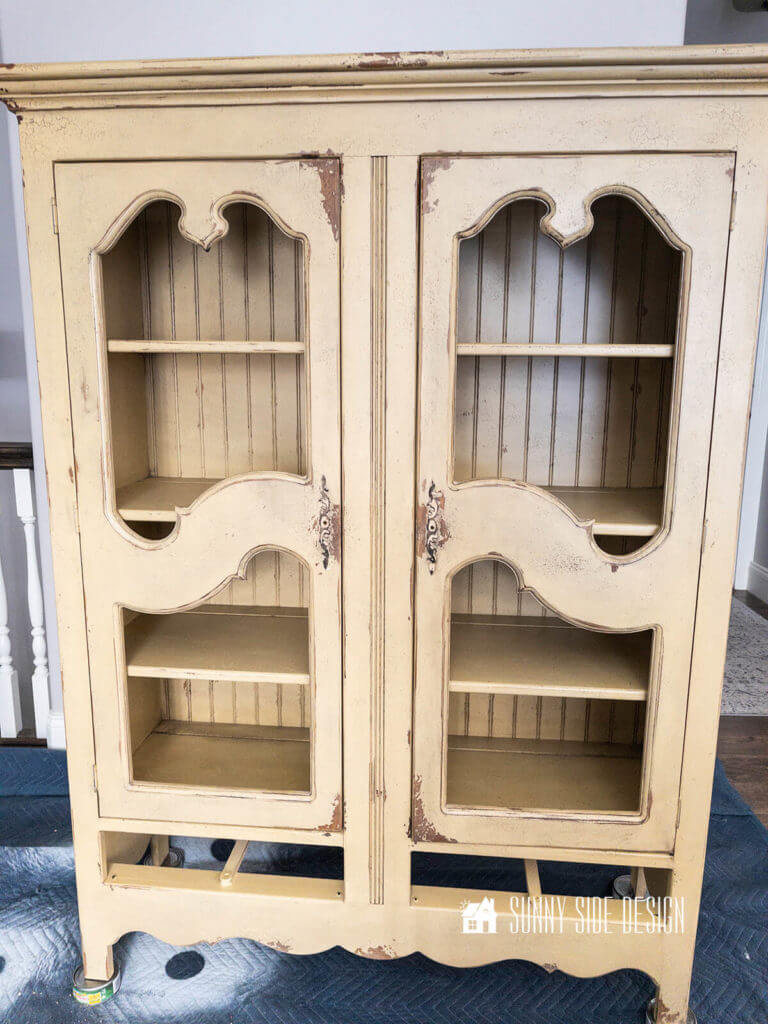 Before photo of the distressed, pale yellow china cabinet with the hardware cloth removed and the drawers removed placed on a blue drop cloth.