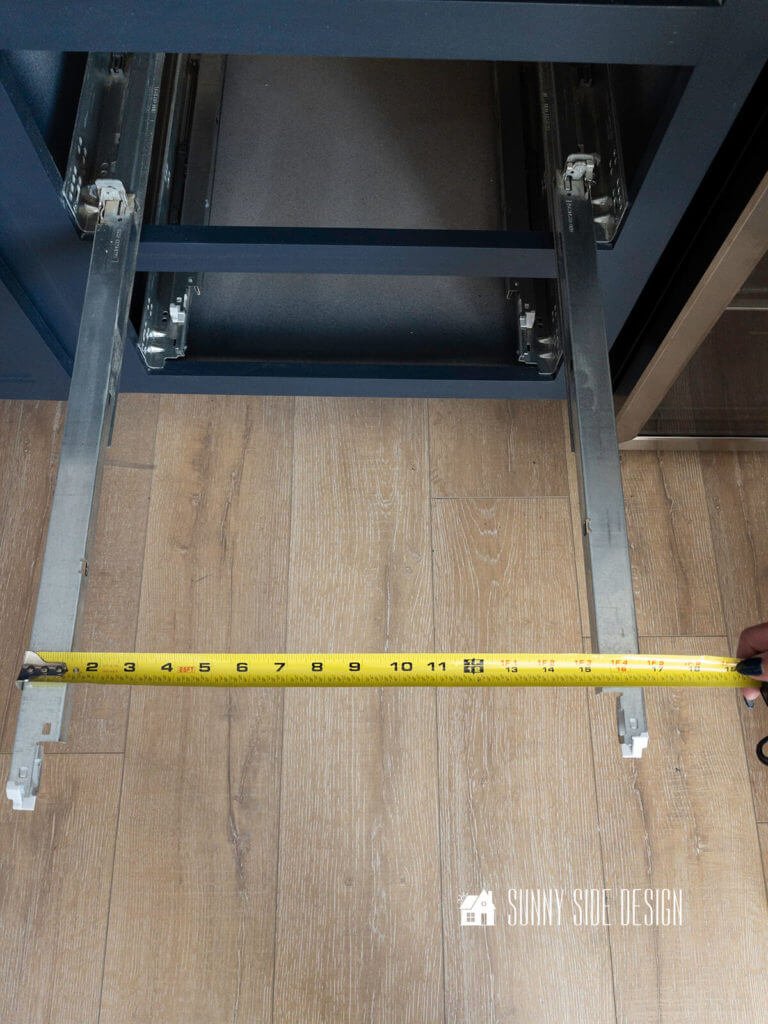 Undermount drawer slide pulled out in a navy blue cabinet box, with a tape measure, measuring the width of the drawer slides.