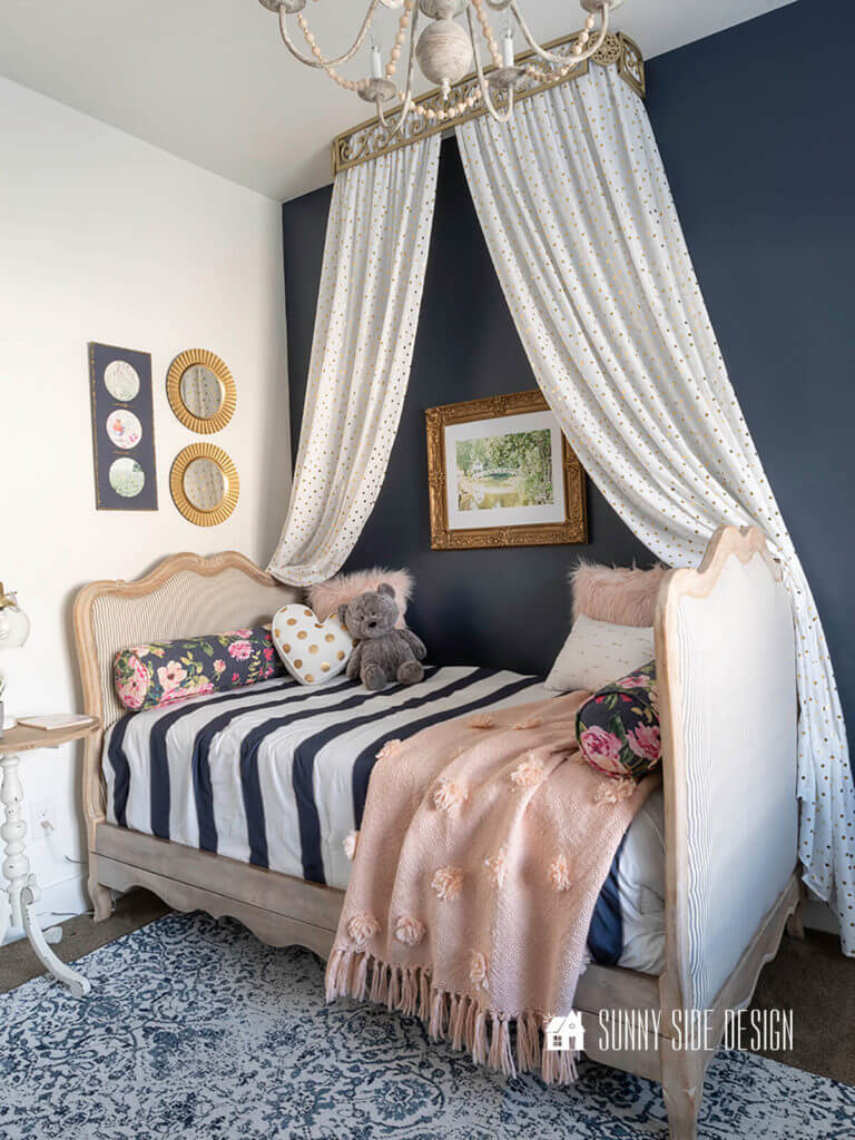 Flea market flip ideas, 2 French provential twin beds are upcycled into a French style daybed with blue and white ticking upholstery and the wood is a natural wood finish. Navy blue and white striped bedding, floral and blush fur pillows. White and gold polka dot form a canopy over the bed, navy blue accent wall, watercolor art on walls.