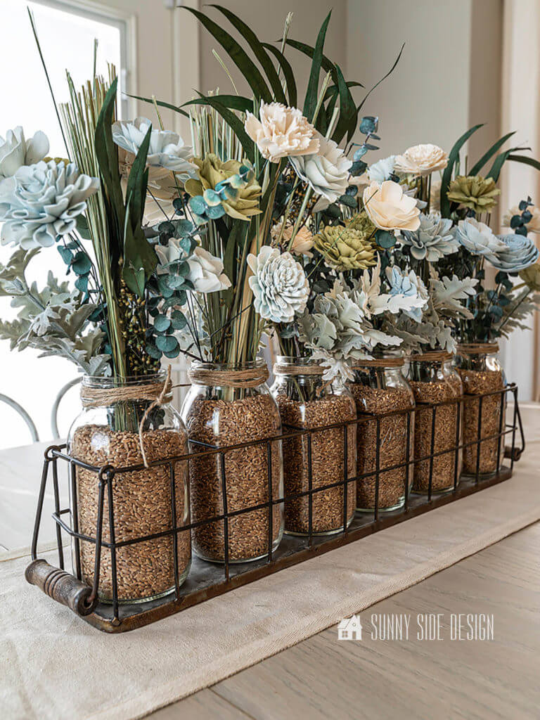 A beautiful Sola wood flower arrangement in glass canning jars with wheat berries in a wire basket on a linen table runner on a natural wood table top.