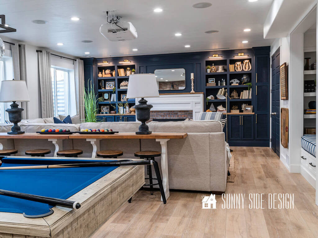 Basement family room ideas, pool table with blue felt, natural wood top and white let sofa table, rustic black lamps with white lampshades, navy blue built ins with white fireplace surround, natural line look drapery.