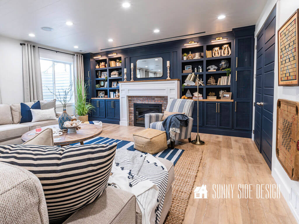 Navy blue bookshelves with a white shaker style fireplace. blue and white stripe chair with tan weave ottoman, brass floor lamp. Vintage games mounted on wall, navy blue closet doors, sisal and blue and white striped rug layered over white oak laminate floor. Linen look curtains, cream colored sectional with blue and white pllow and a natural wood coffee table.
