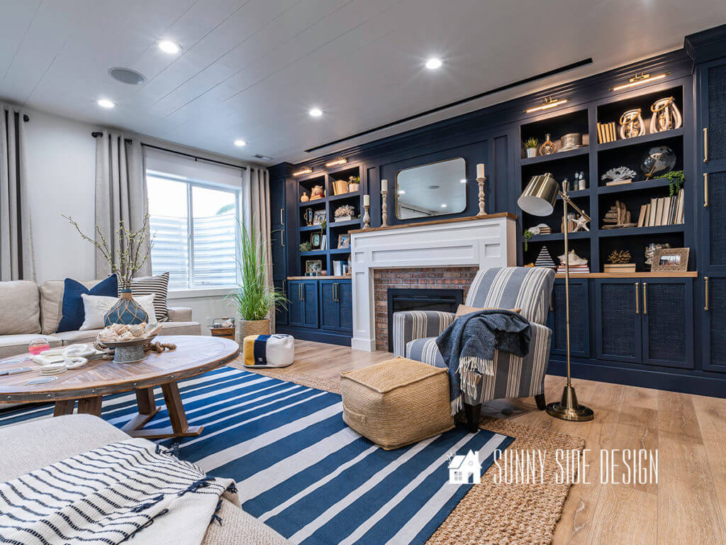 Basement family room ideas, cream colored sectional with blue and white pillows, sisal and blue and white striped rug on floor with natural wood coffee table, blue throw blanket on blue and white striped chair, rustic table lamps on sofa tables, floor plants, linen look drapes, navy blue built in bookshelves around a shaker style fireplace.
