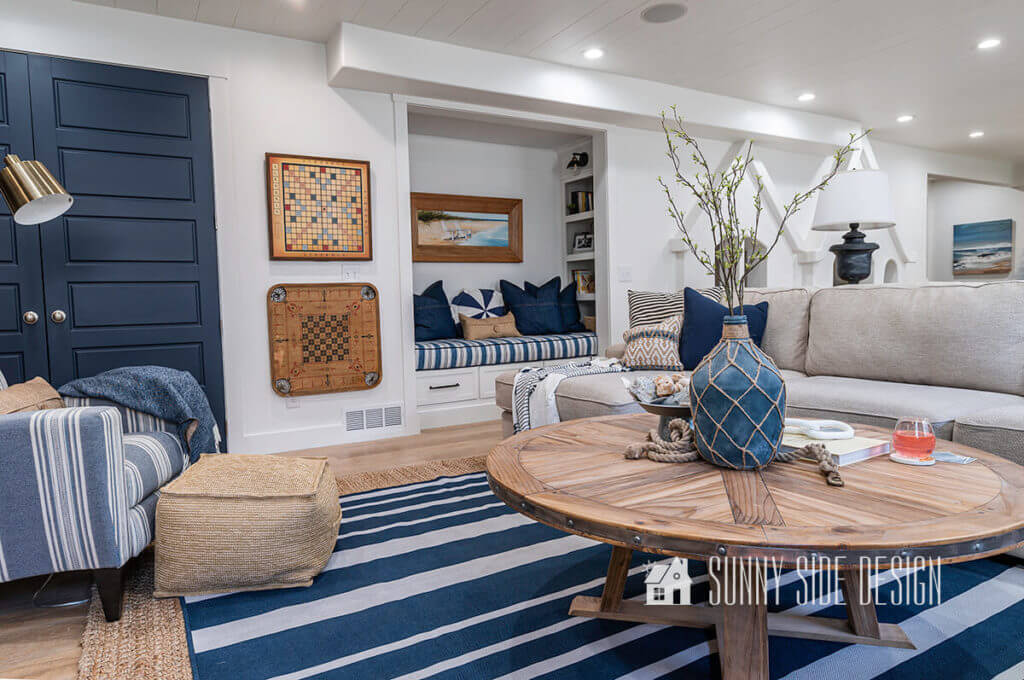 Basement family room ideas, cream colored sectional with blue and white pillows, sisal and blue and white striped rug on floor with natural wood coffee table, blue throw blanket on blue and white striped chair, reading nook, vintage games mounted to wall.