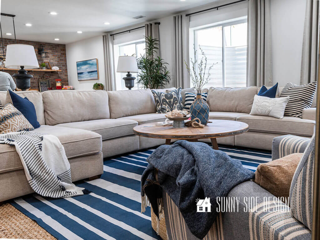 Basement family room ideas, cream colored sectional with blue and white pillows, sisal and blue and white striped rug on floor with natural wood coffee table, blue throw blanket on blue and white striped chair, rustic table lamps on sofa tables, floor plants, linen look drapes on 3 windows.