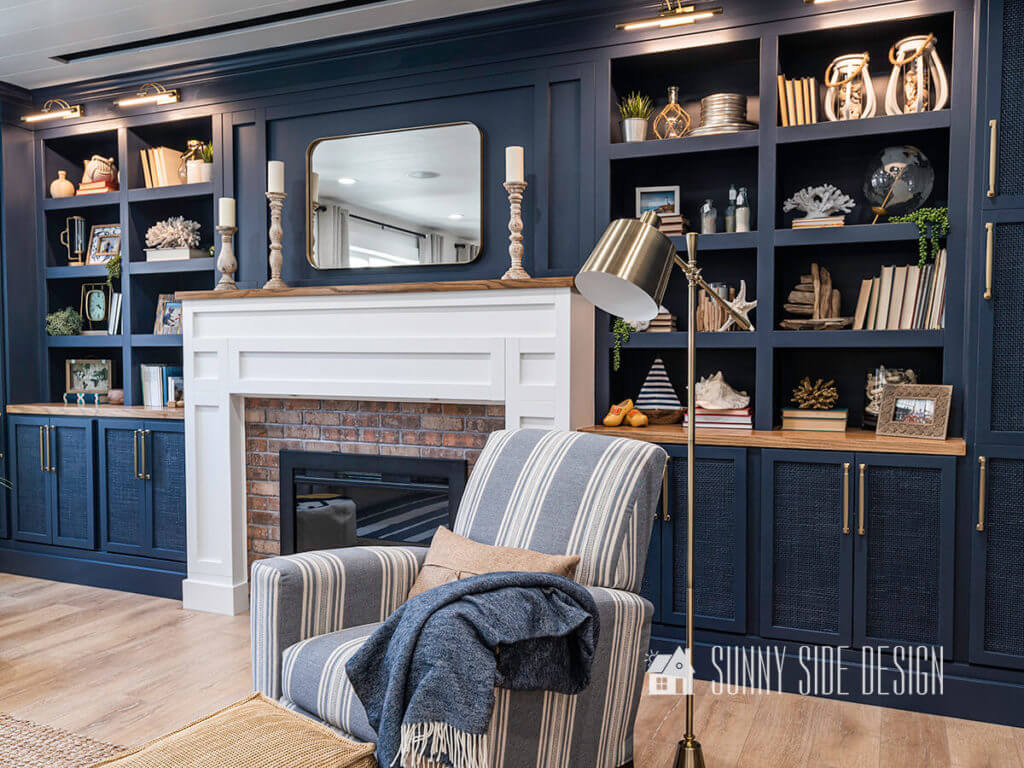 Basement family room ideas, blue and white striped chair with blue throw, brass floor lamp. Navy blue built in bookshelves with a white shaker style fireplace surround, wood mantle decorated with rustic candlesticks.