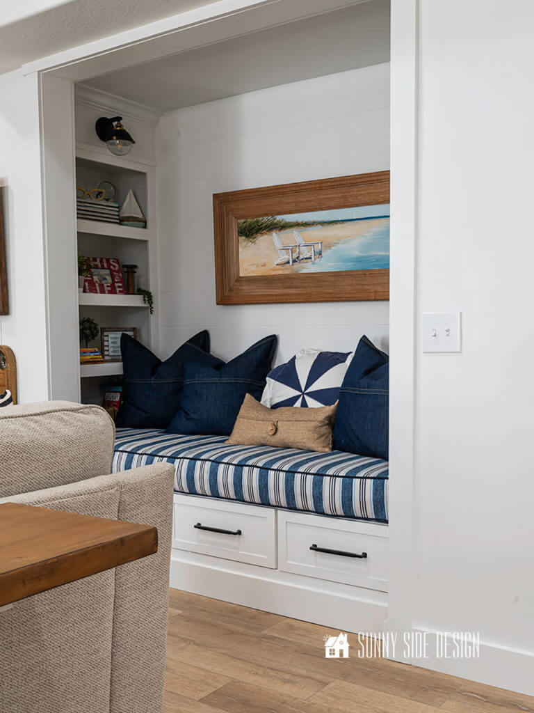 Basement family room ideas, left view of reading nook, blue and white cushion, white bookshelves decorated with photo grames, books, sailboat and plants.