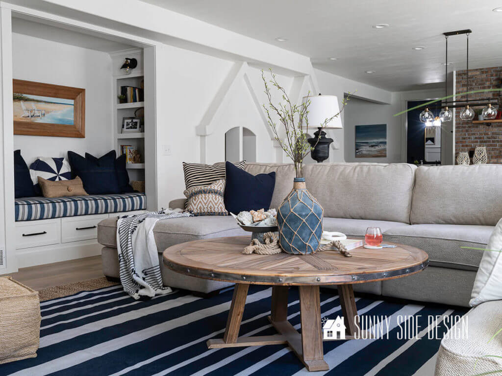 Basement family room ideas, blue and white striped rug layered over a sisal rug, natural wood round coffee table with a blue jug with green stems, cream color sectional sofa with blue and white pillows. Built into the wall is a reading nook with a blue and white striped cushion with pilllows.
