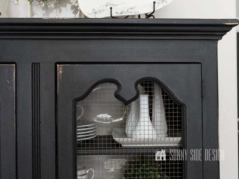 Close up view of black chalk painted china cabinet with hardware cloth in doors filled with a variety white dishes and vases.