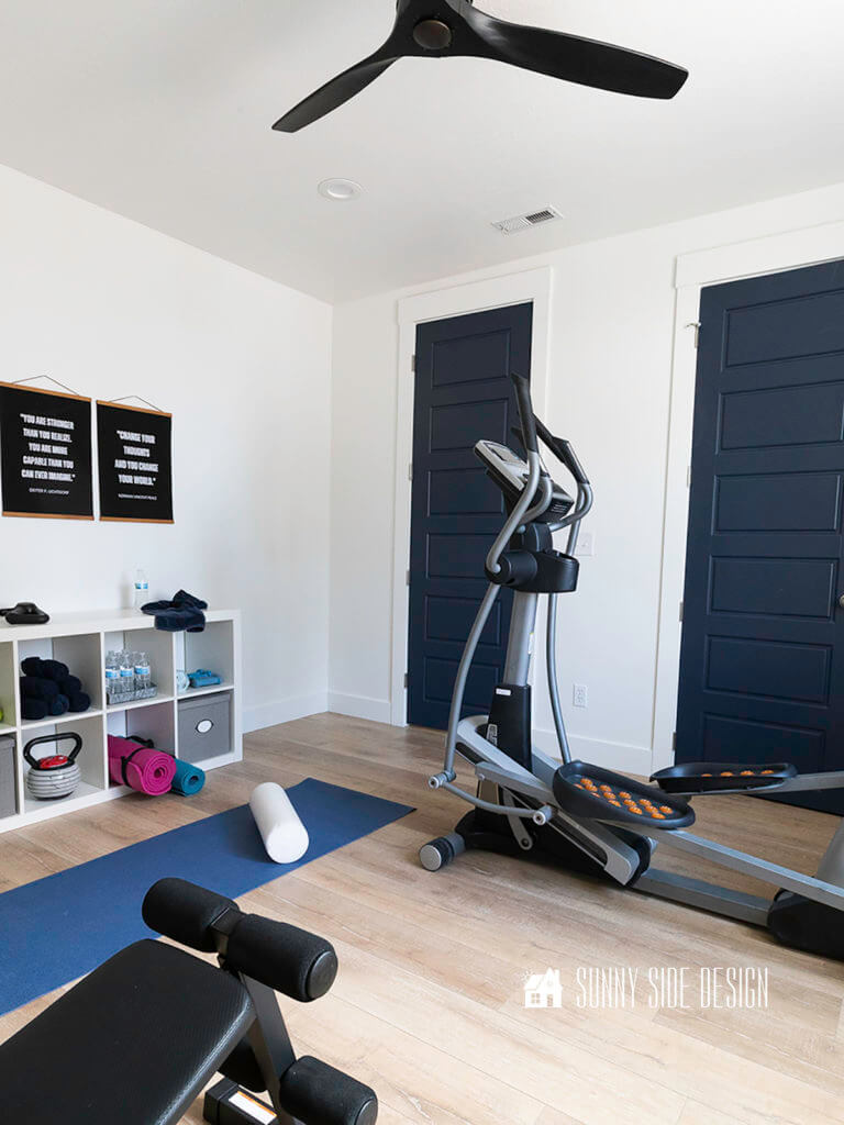 Painting a room, finished white painted room with shelves for storing home gym equipments and black and white motivational text art on the wall, eliptical machine and a blue yoga mat with a foam roller on the wood floor. Navy blue doors in the white painted room.