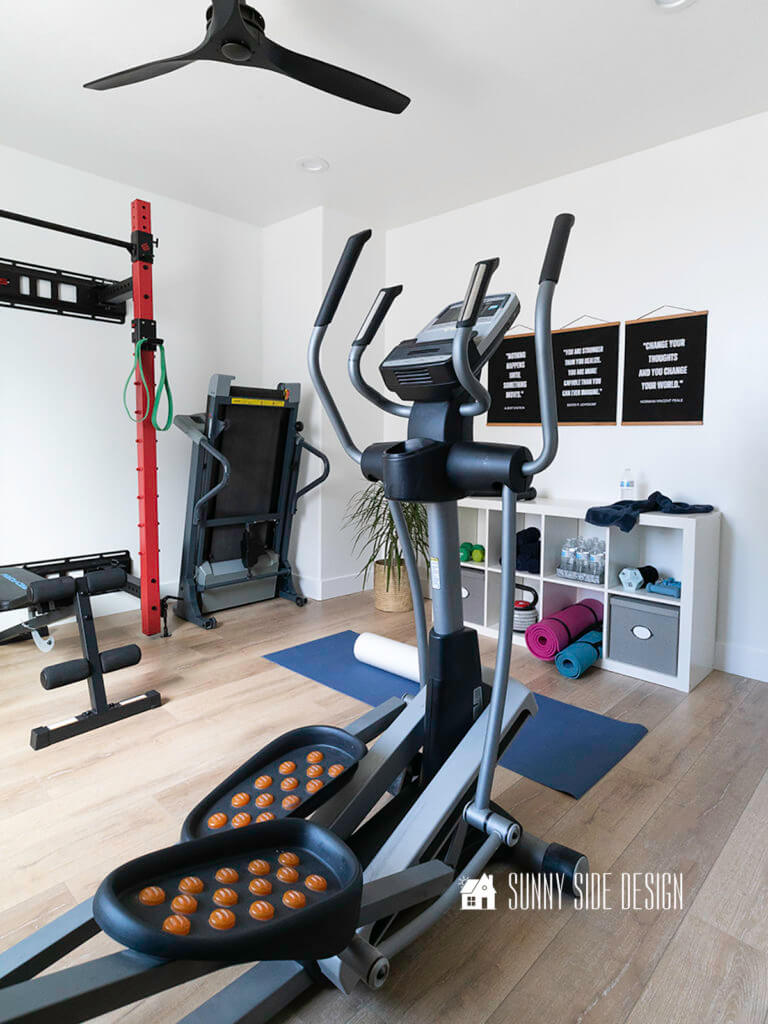 Painting a room, finished white painted room with shelves for storing home gym equipments and black and white motivational text art on the wall, green plant in the corner of the room, eliptical maching, treadmill, black ceiling fan.