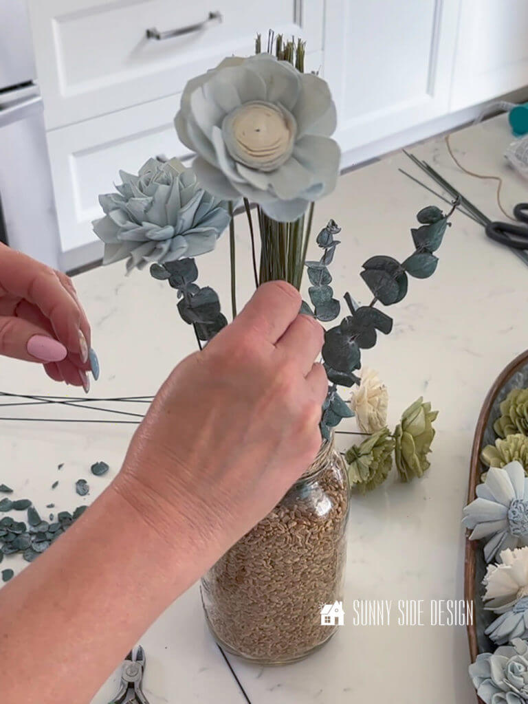 Woman's hand placing a blue dyed Sola wood flower into the floral arrangment in a glass jar.
