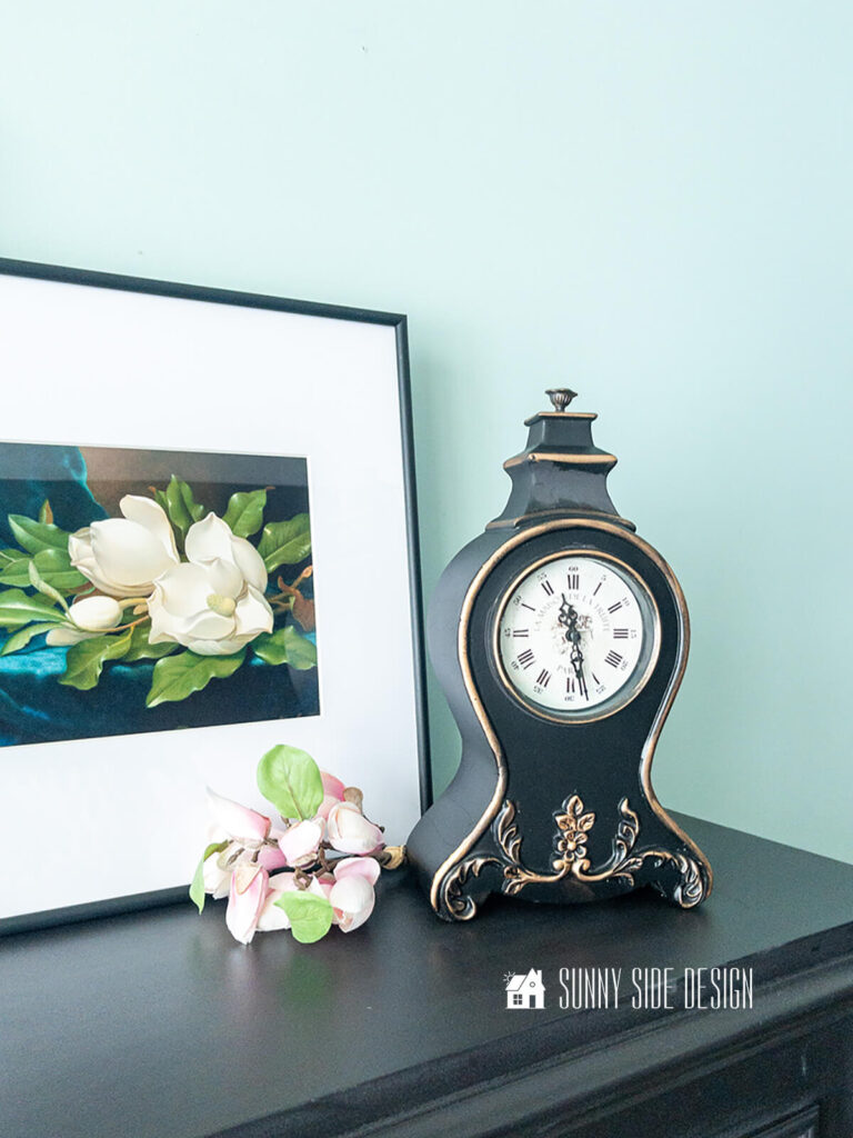 Pretty French inspired clock upcycled with black paint and gold Rub n Buff, styled on a black cabinet with floral art and a floral bouquet.
