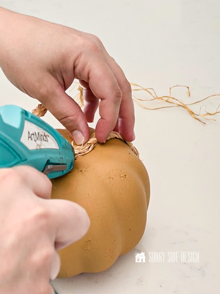 Woman applying hot glue to the loose ends of the raffia on the foam pumpkin.