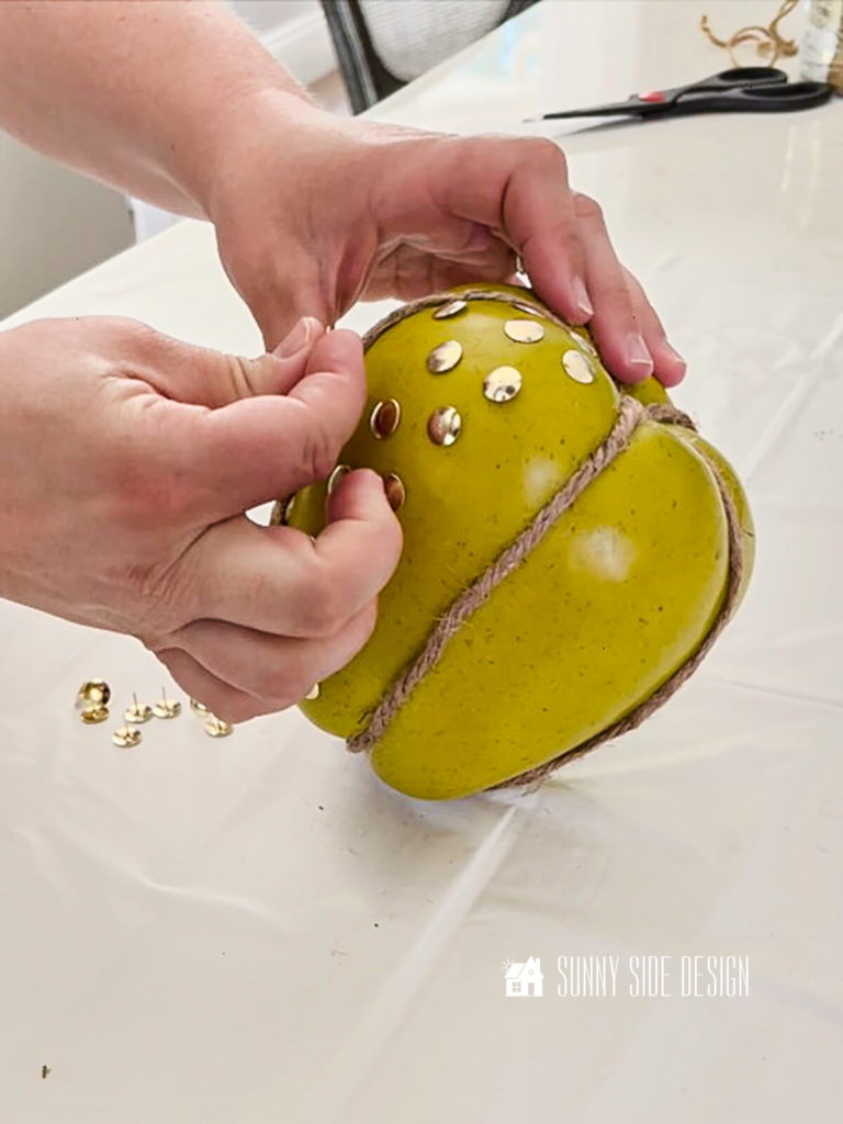 Woman's hand pressing in gold thumbtacks into a large foam apple for the next pumpkin DIY.