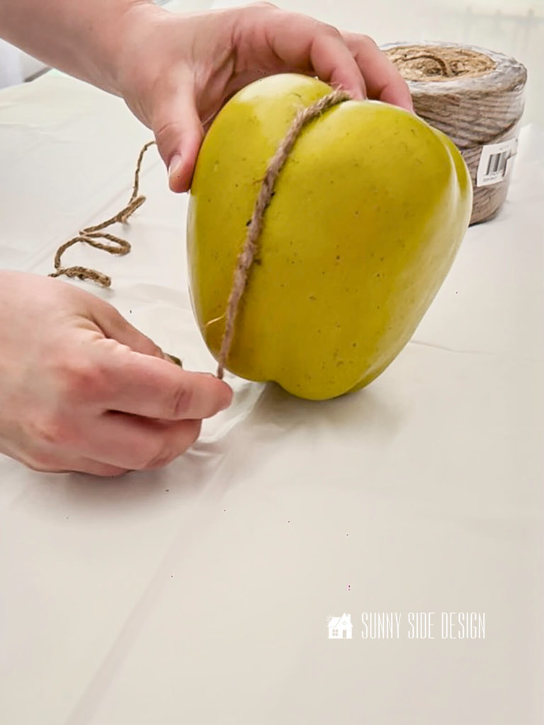 Woman wraps twine into the grooves of a large faux apple to form sections for the pumpkin DIY.