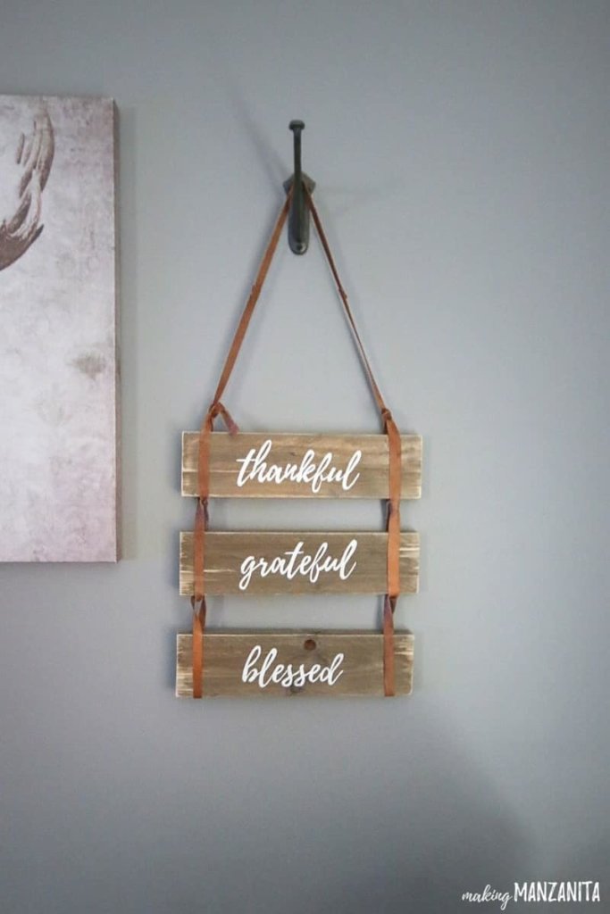 DIY Fall Decor ideas, 3 pieces of brown painted pallet wood with the words "thankful, grateful, blessed" stenciled on with white paint. The pallet wood boards are tied together with strips of leather. The sign is hanging on a wall hook agains a grey wall.