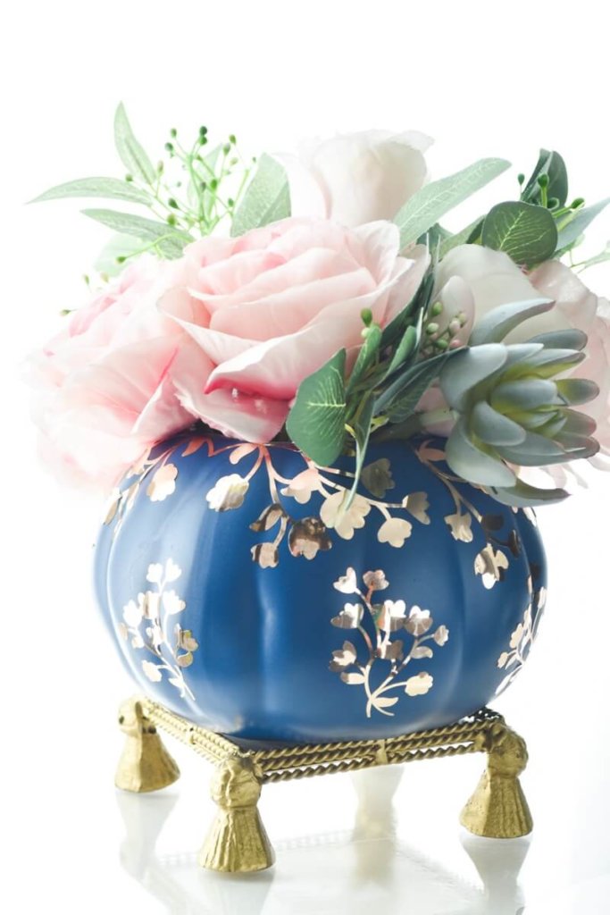 Navy blue painted pumpkin with gold chinoiserie design applied with vinyl, setting on a gold stand with pink and white flowers.