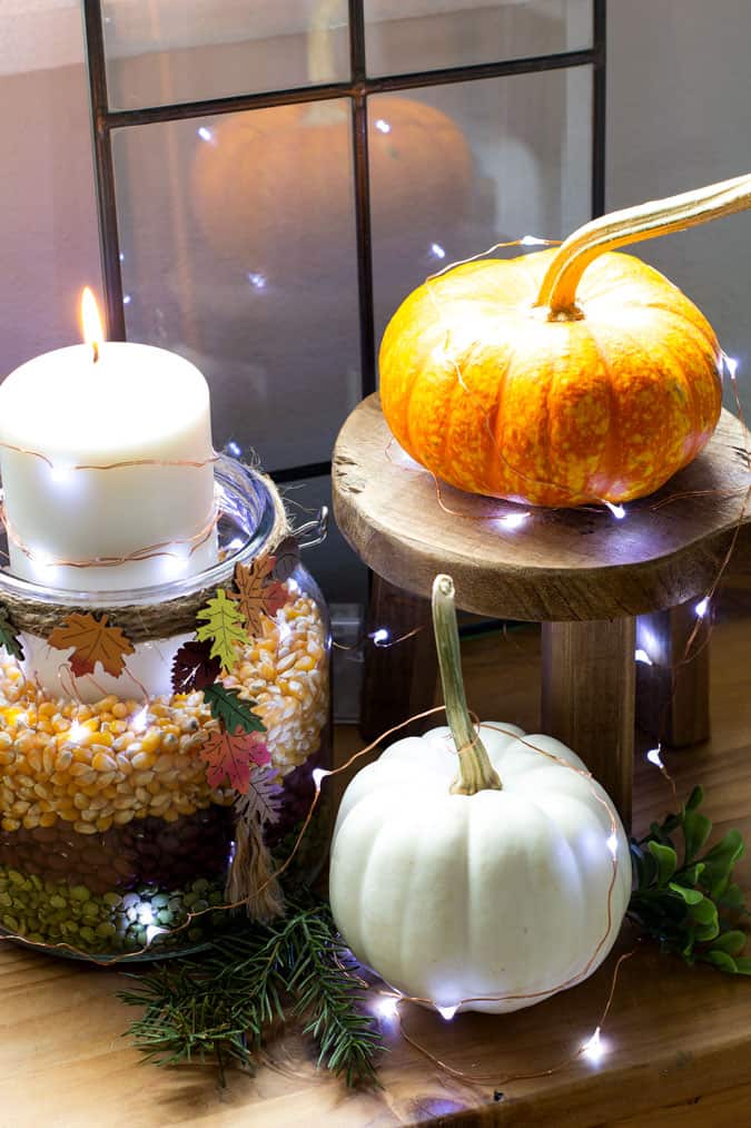 Fairly lights arranged around mini pupkins, wood stand, and jar filled with assorted grains and a candle.