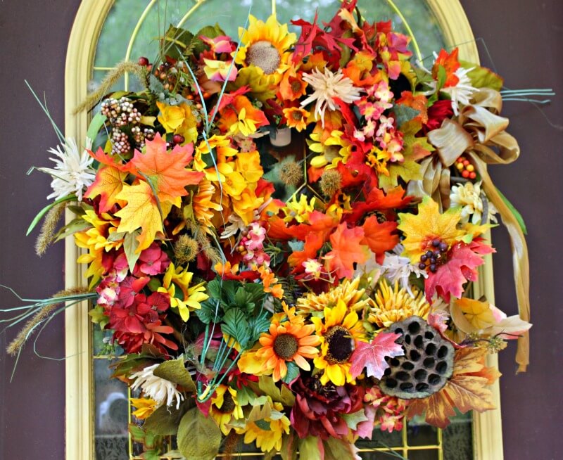 Fall leaf wreath with sunflowers, seed pods, grasses, mums and ribbon on a brown door.