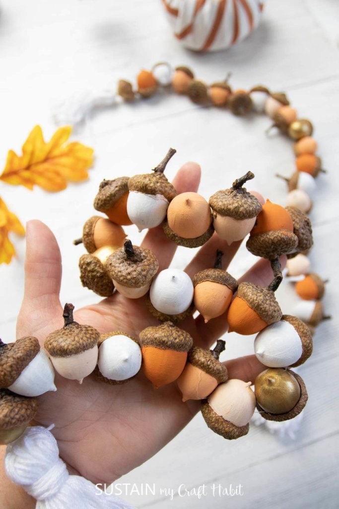 Woman's hand holding a DIY acorn garland. Natural acorns painted white, cream, coral and orange.