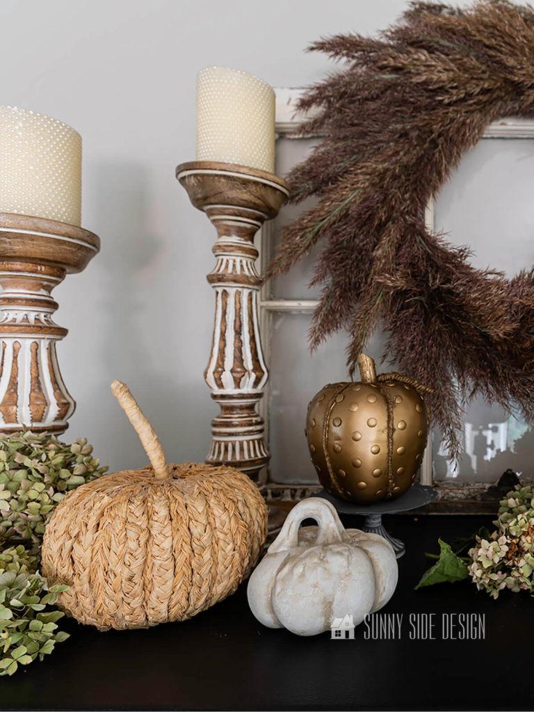 Braided raffia pumpkin setting on a black cabinet with 2 natural wood candlesticks, dried hydrangeas, pampas grass wreath along with a gold and ceremic pumpkin.