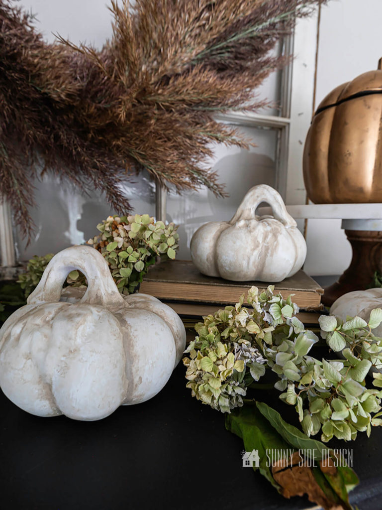 Pumpkin DIY, thrift store ceramic pumpkins painted with a farmhouse lookl white and shadded with greige paint, dried hydrangeas ane one pumpkin elevated on a stack of vintage book. Pampas grass wreath hung on a vintage window.