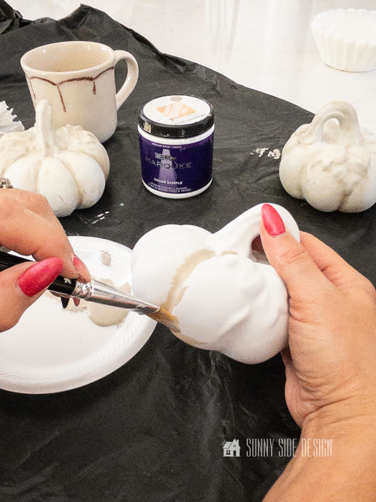 Woman applies taupe paint to a white painted thrift store pumpkin.