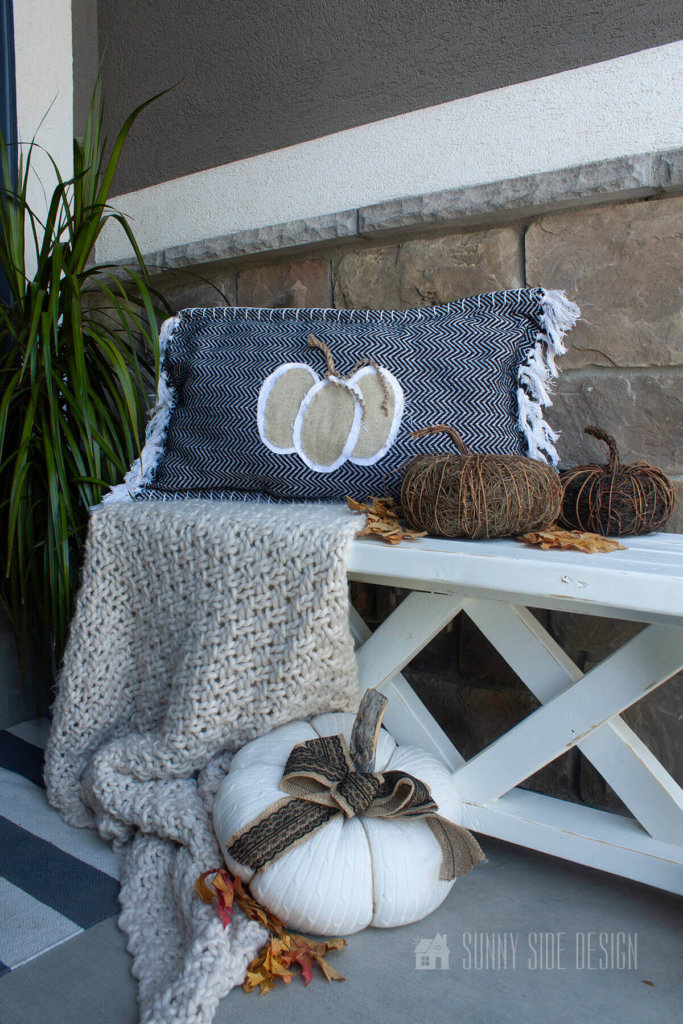 DIY fall pumpkin pillow with natural linen and rope, made from a dollar store rug. Setting on a white farmhouse style bench with a throw blanket and grapevine pumpkins and maple leaves.