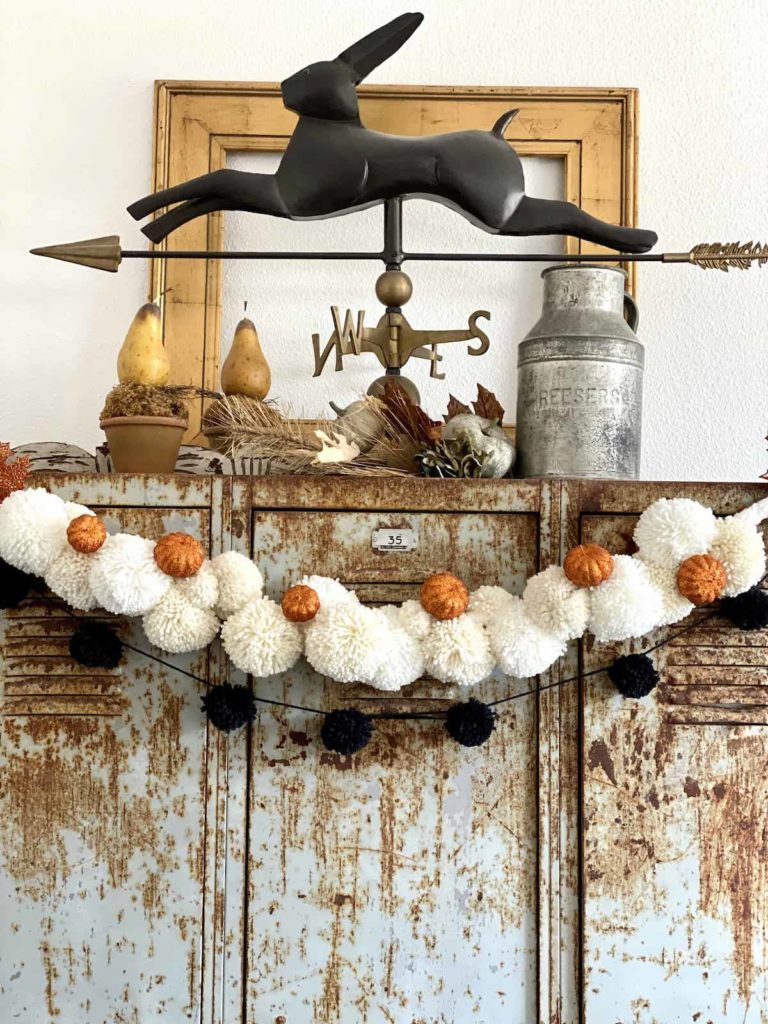 Easy DIY Halloween pom pom garland hanging on rusty metal lockers. a bunny weathervane sits o top with pear topiaries and a metal jug.