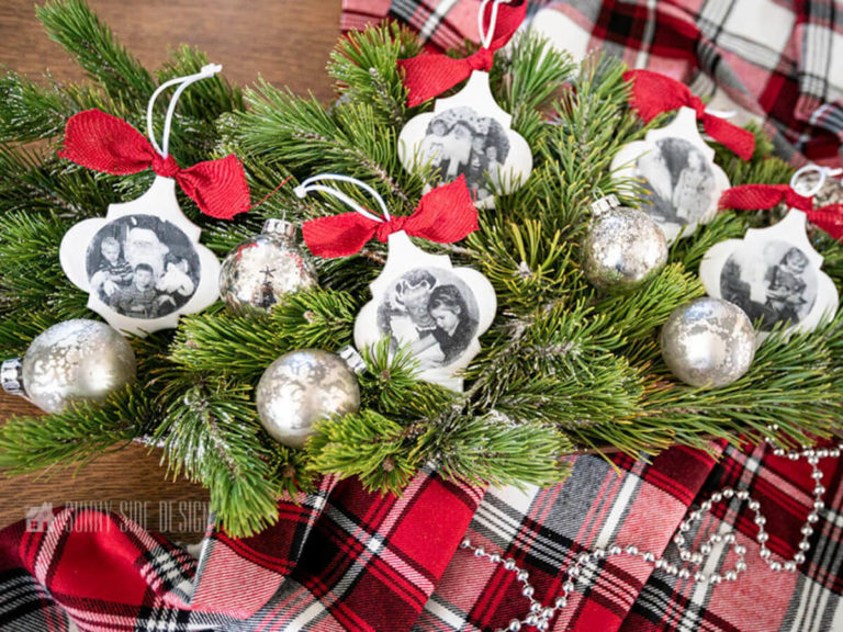 Personalized Christmas ornaments made with white arabeque tiles and old black and white photos placed in a bed of fresh greenery and mercury glass ornaments with a red plaid flannel cloth.
