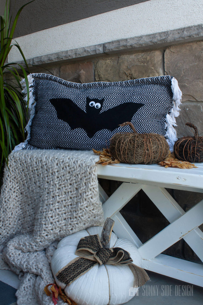 Black and white bat pillow with a ivory knit throw blanket, sweater pumpkin and natural vine pumpkins on a white farmhouse bench and easy Halloween decor ideas for your home.