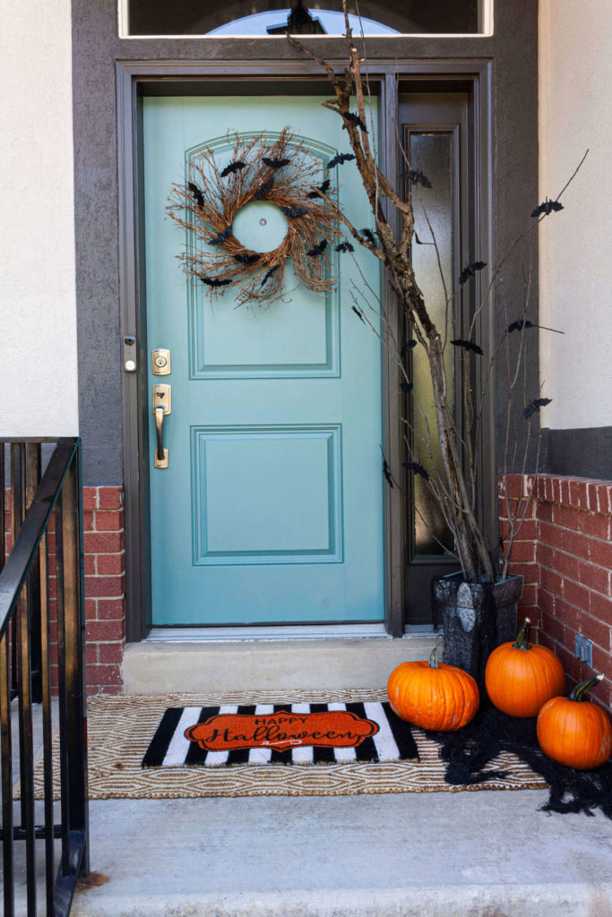 Light blue front door with a twig and bat wreath, a large branch is placed in a galvanized metal container and bats are hanging from the branches. Black orange and white welcome mat layered with a jute rug and orange pumpkins placed on black creepy cloth.