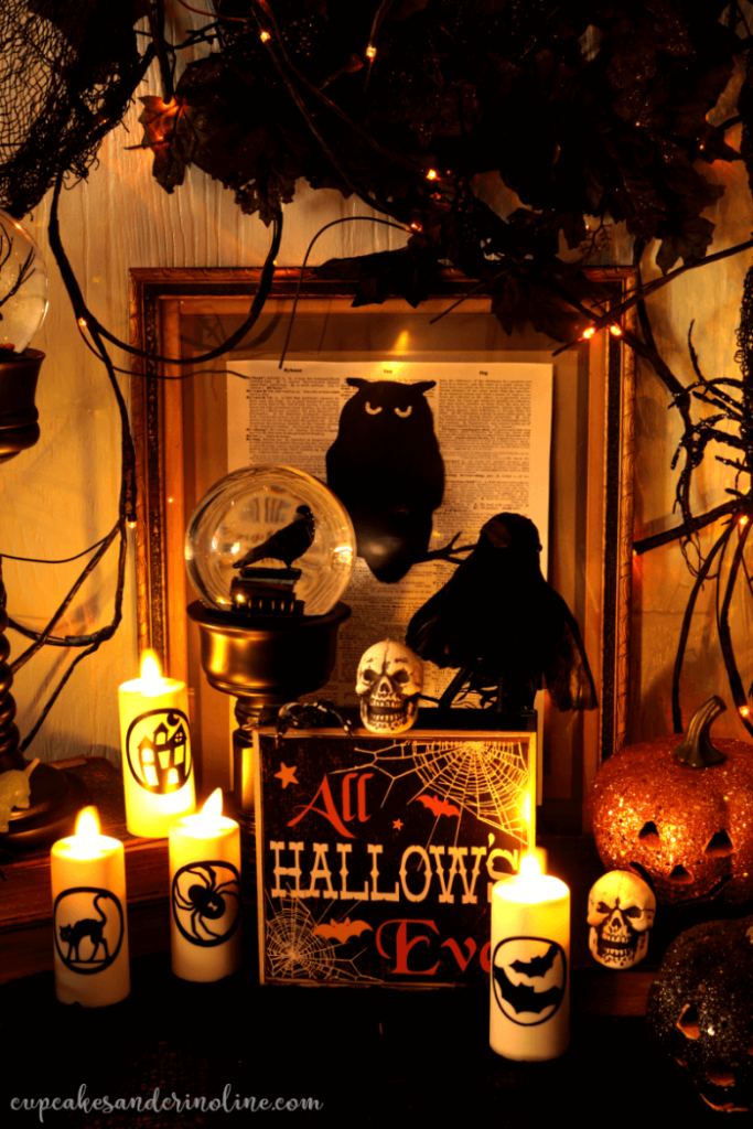 Lit Halloween candles with a candle stick with a fortune tellers ball, mini skulls, and pumpkins, and framed book page art with an owl. Twig, mini lights and black flowers add to the ambiance.